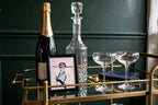 Styled lifestyle photo — the small Art Deco lady framed artwork (9.5 × 11.5 in) standing on a brass bar cart beside a champagne bottle and decanter (display inspiration)
