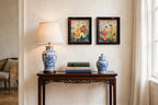 Styled lifestyle photo — the small framed Asian textile pair (17 × 14 in each) above a dark-wood side table with a porcelain lamp (display inspiration)