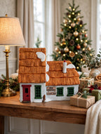 Styled lifestyle photo — the Cratchit cottage on a console table beside a lamp and wrapped gifts, Christmas tree behind (display inspiration)