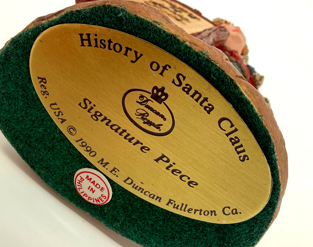 Close-up of the underside gold label — History of Santa Claus, Signature Piece, Duncan Royale, Reg. USA © 1990 M. E. Duncan Fullerton Ca., with the Made in Philippines sticker on the green felt base