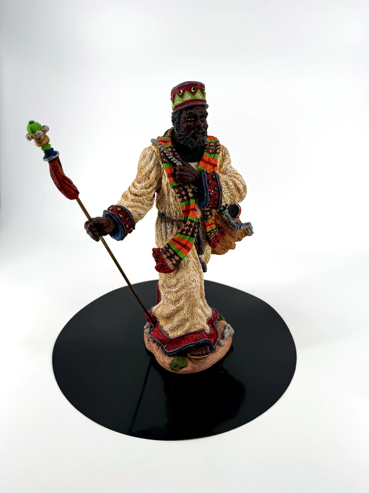 African American Santa figurine front view on a black display disc — cream robe, African-pattern scarf, jeweled crown, staff with red tassel, and gift sack