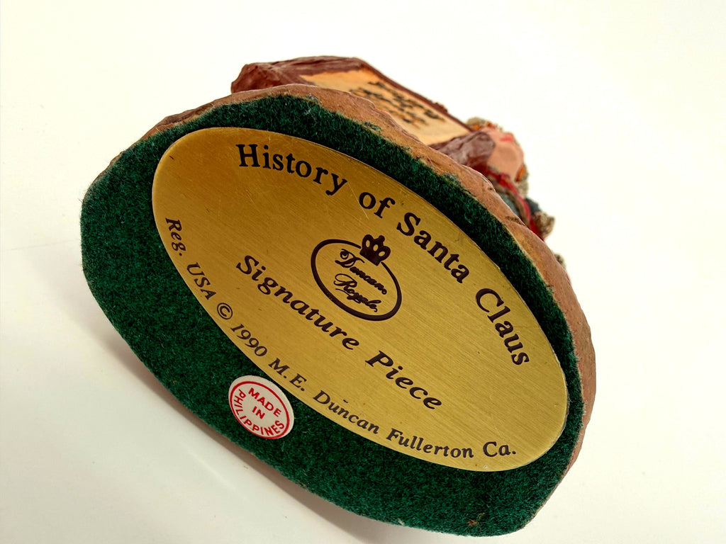 Underside of the Signature Piece showing the original gold label: History of Santa Claus - Signature Piece - Duncan Royale - Reg. USA 1990 M. E. Duncan Fullerton Ca., with the Made in Philippines sticker, on the green felt base
