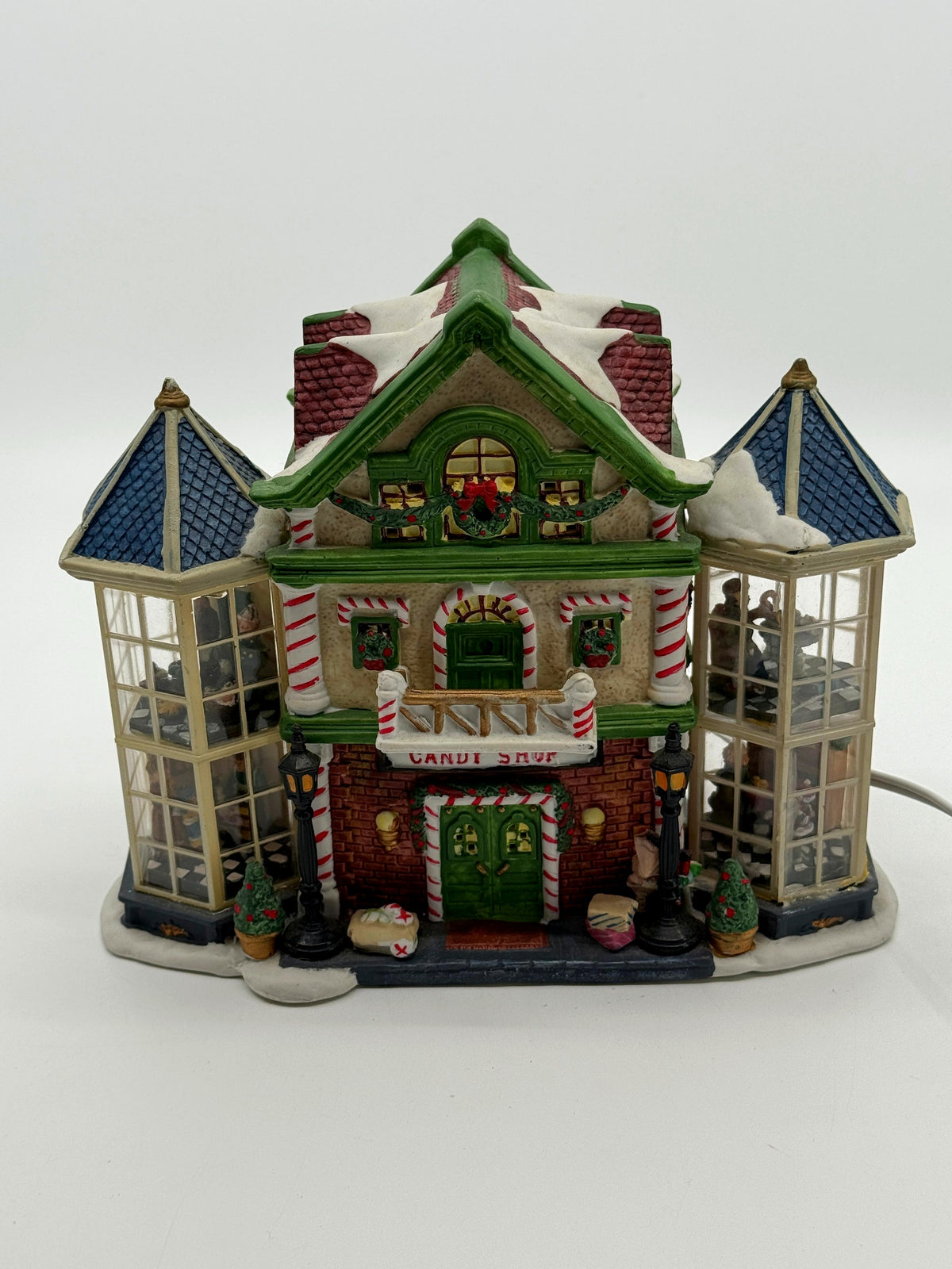 Front view of the Child's Wonder Candy Shop village building with snow-covered roofs, wreath attic window, and lampposts at the green double doors