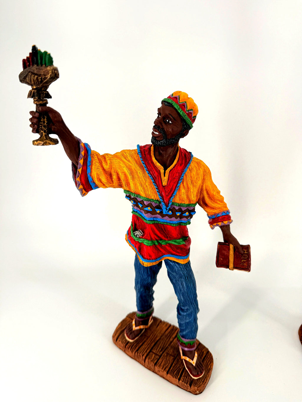 The male Kwanzaa figure alone — kinara raised in one hand, book in the other, colorful patterned tunic and cap