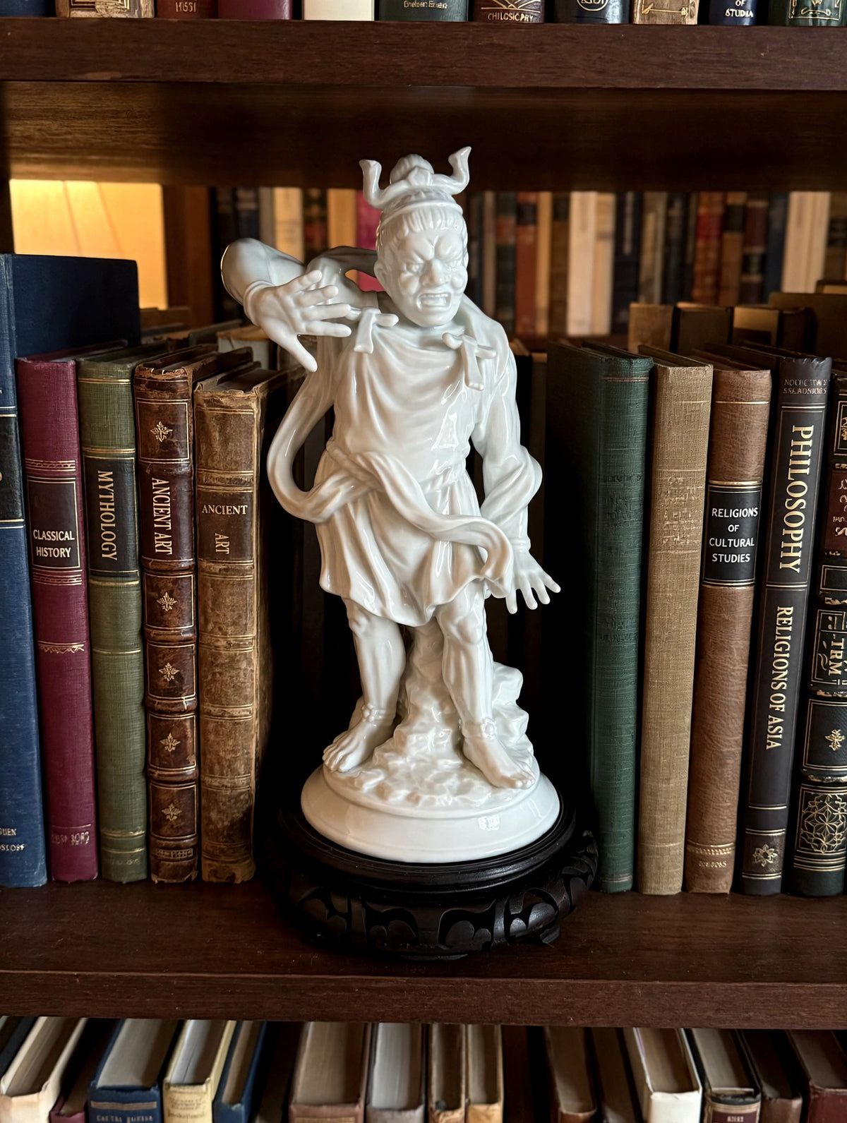 Styled lifestyle photo — the white porcelain warrior guardian figurine (about 11–12 in tall on its stand) on a bookshelf among hardcover books (display inspiration)