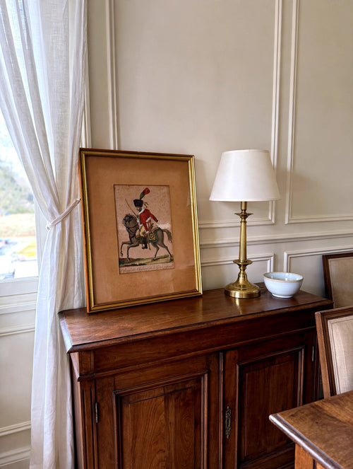 Styled lifestyle photo — the framed Martinet chasseur engraving leaning on a dark wood sideboard in a bright dining room (display inspiration)