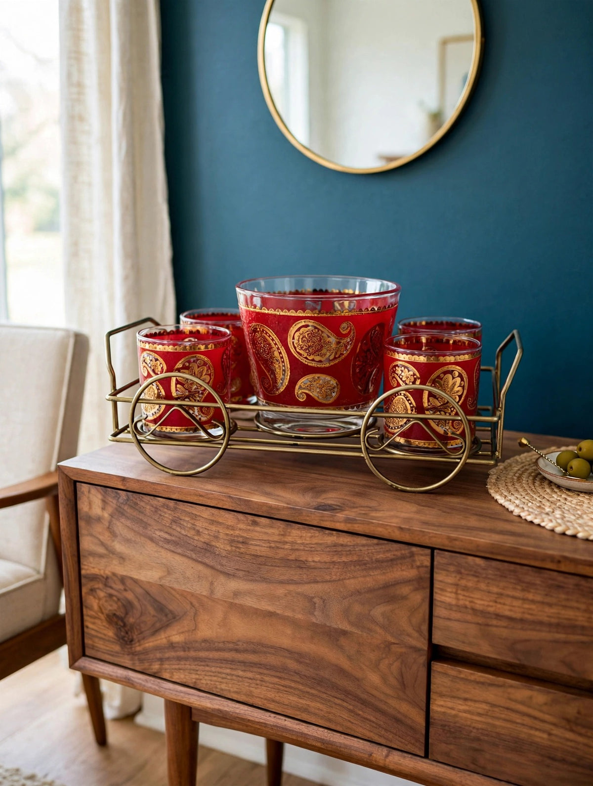 Styled lifestyle photo — the Culver red and gold paisley barware set in its brass caddy on a walnut sideboard in a bright mid-century bar nook with deep teal walls (display inspiration)