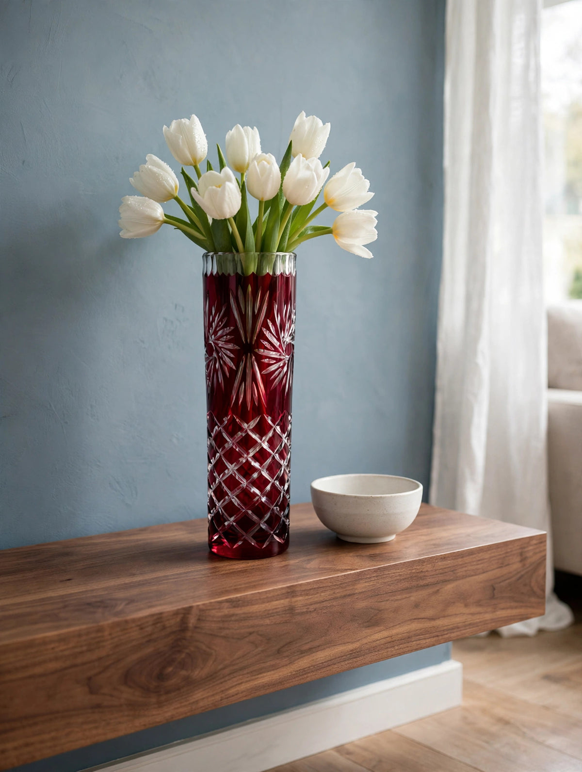Styled lifestyle photo — the ruby cut-to-clear vase with white tulips on a walnut console table in a bright living room with dusty blue walls (display inspiration)