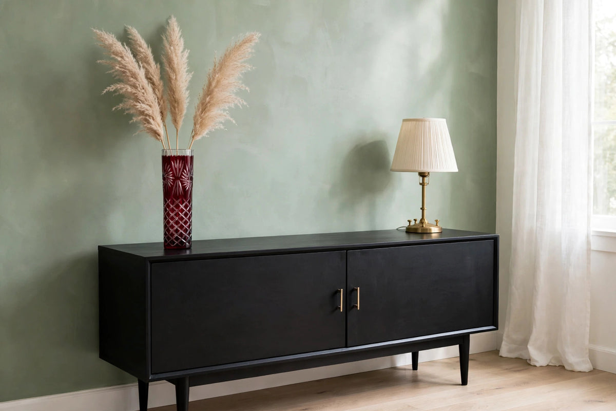 Styled lifestyle photo — the ruby cut-to-clear vase with pampas grass on a black mid-century sideboard in a bright room with sage green walls (display inspiration)