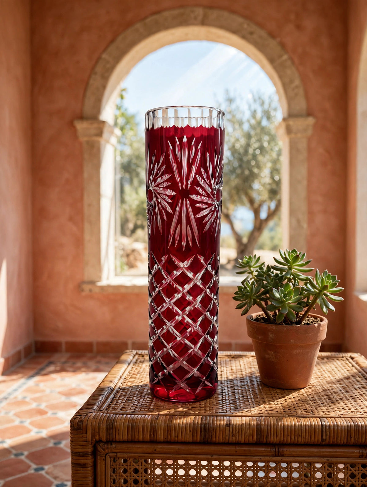 Styled lifestyle photo — the ruby cut-to-clear vase on a rattan console table in a bright Mediterranean sunroom with terracotta walls (display inspiration)