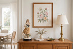 Styled lifestyle photo — the Armani "Roses of May" figurine on a wood sideboard beneath a framed floral print in a bright dining room (display inspiration)