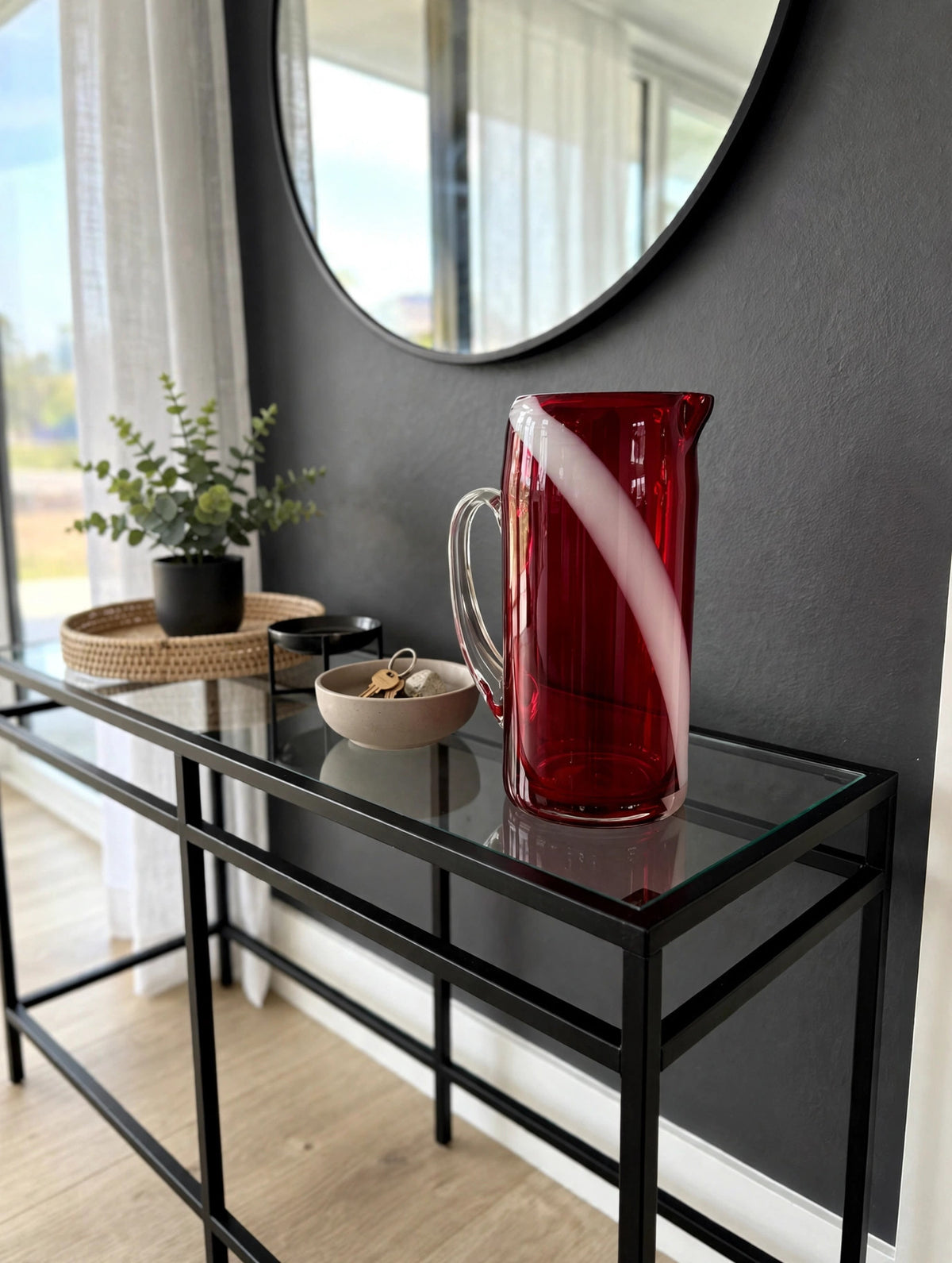 Styled lifestyle photo — the ruby swirl glass pitcher on a black metal and glass console table in a bright entryway with a charcoal accent wall (display inspiration)