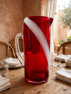 Styled lifestyle photo — the ruby swirl glass pitcher on a rustic dining table in a bright Mediterranean dining room with terracotta walls (display inspiration)