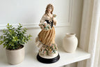 Styled lifestyle photo — the Armani "Roses of May" figurine on a white shelf beside a potted plant in a bright room (display inspiration)
