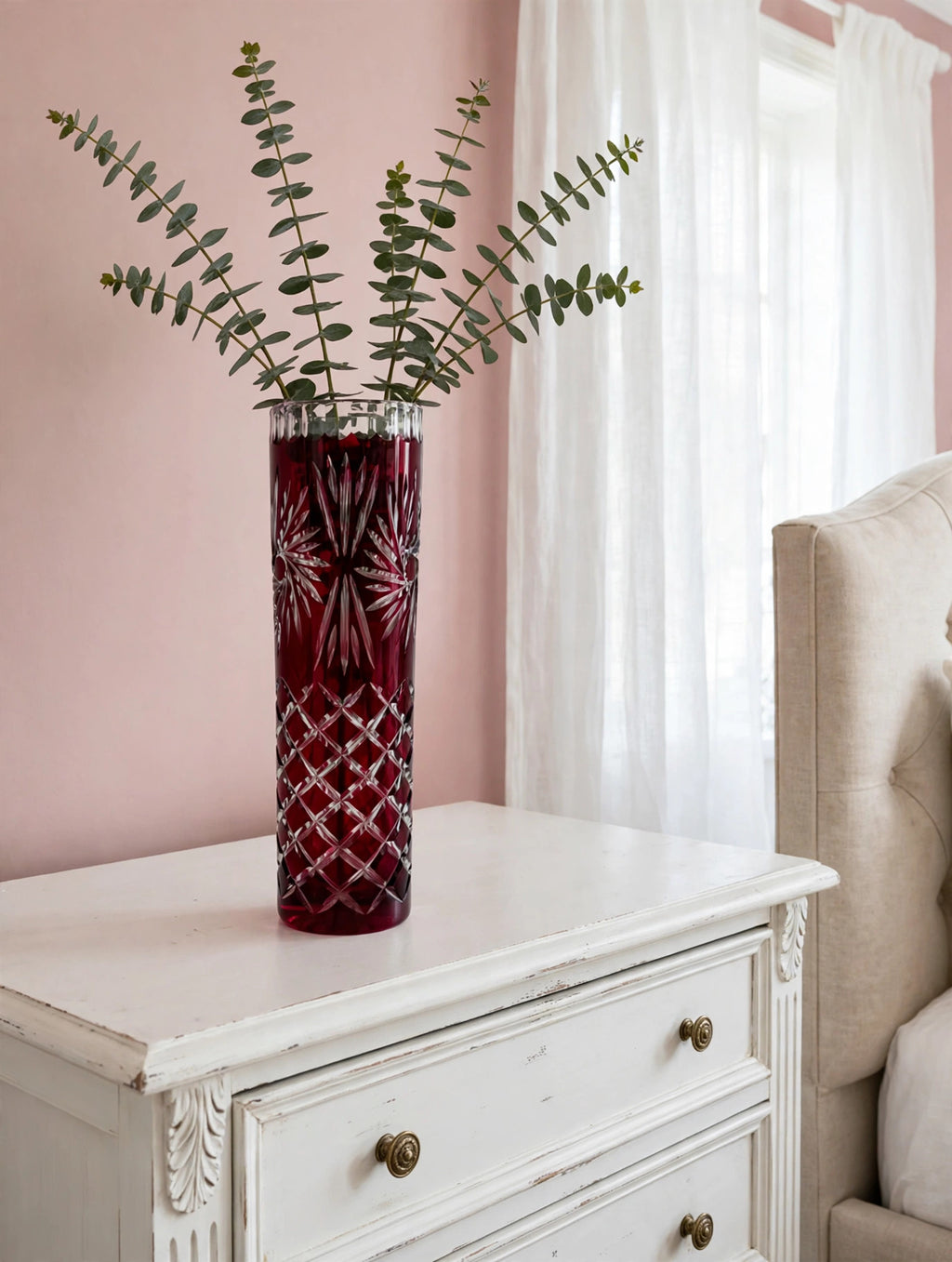 Styled lifestyle photo — the ruby cut-to-clear vase with eucalyptus on a white dresser in a bright bedroom with blush walls (display inspiration)