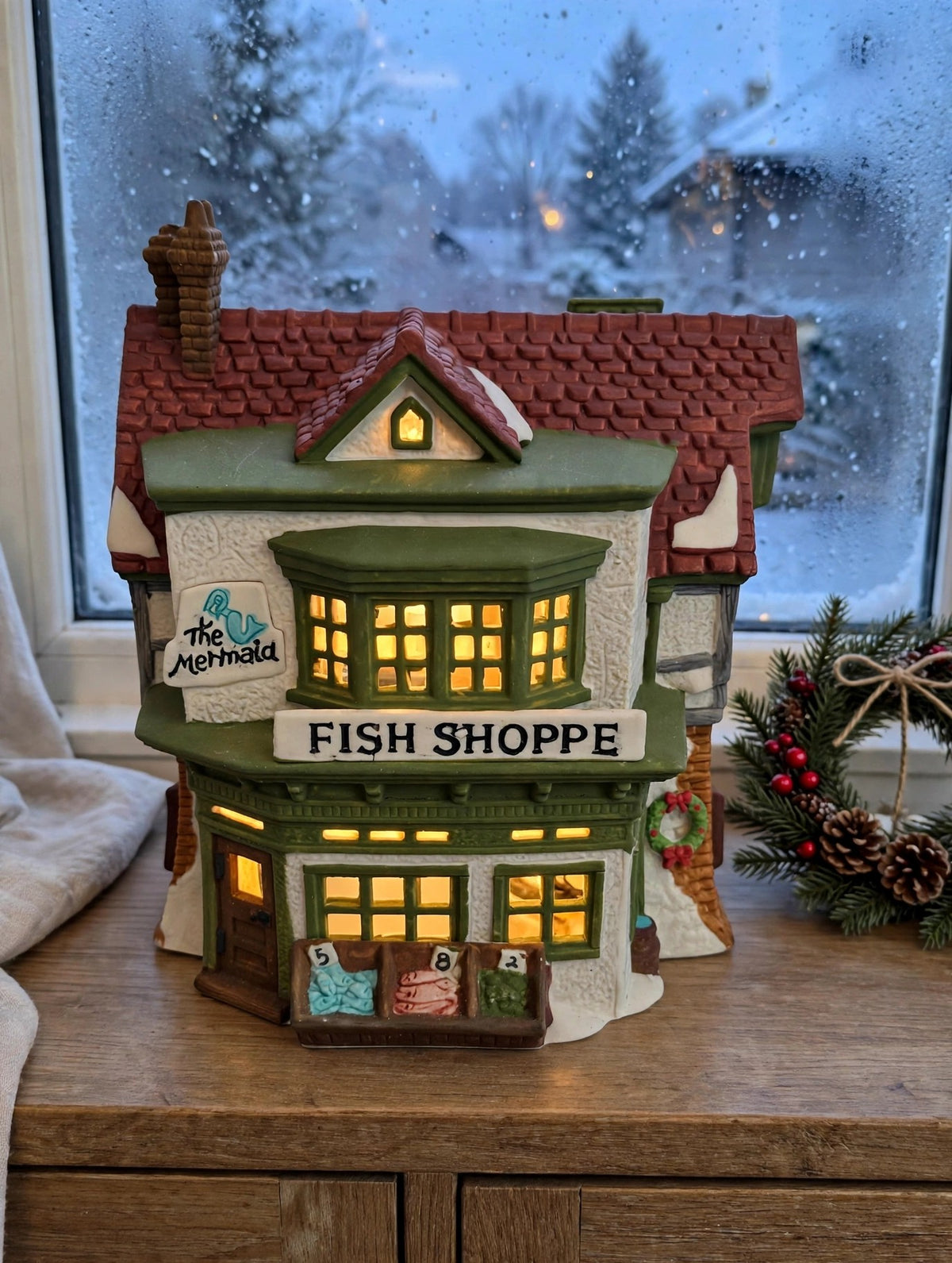 Department 56 Dickens' Village The Mermaid Fish Shoppe #5926-9 — styled scene: lit on a wooden sideboard by a frosted window at dusk (display inspiration)
