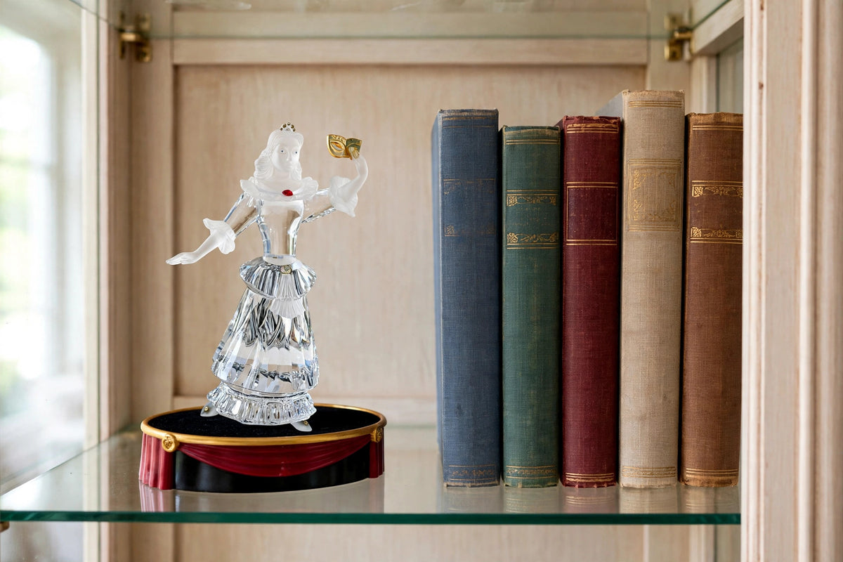 Styled lifestyle photo — the Columbine crystal figurine on its theatrical display stage on a glass curio cabinet shelf beside tall books (display inspiration)