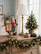 Styled lifestyle photo — the Signature Piece on a vintage wooden sideboard beside a tall brass table lamp, a small tabletop Christmas tree, wrapped gifts and a lit garland (display inspiration)