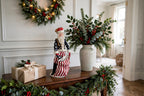 Styled lifestyle photo — on a dark wood entry table beside a tall vase of holly and greenery, with a lit wreath, garland and a wrapped gift (display inspiration)
