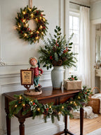 Styled lifestyle photo — the Signature Piece on a dark-wood entry table beside a tall vase of holly and greenery, with a lit wreath, garland and a wrapped gift (display inspiration)