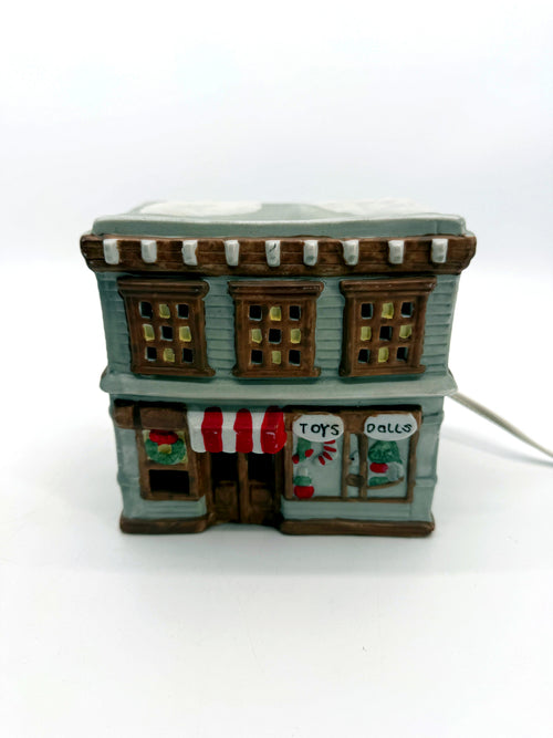 Ceramic Christmas village Toy and Doll shop, front view - hand-painted gray storefront with red and white striped awning, wreath and snow-covered roof
