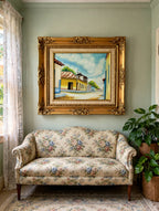 Styled lifestyle photo — the 1984 signed Venezuelan street scene oil shown in a sunlit vintage parlor above a floral settee (display inspiration)