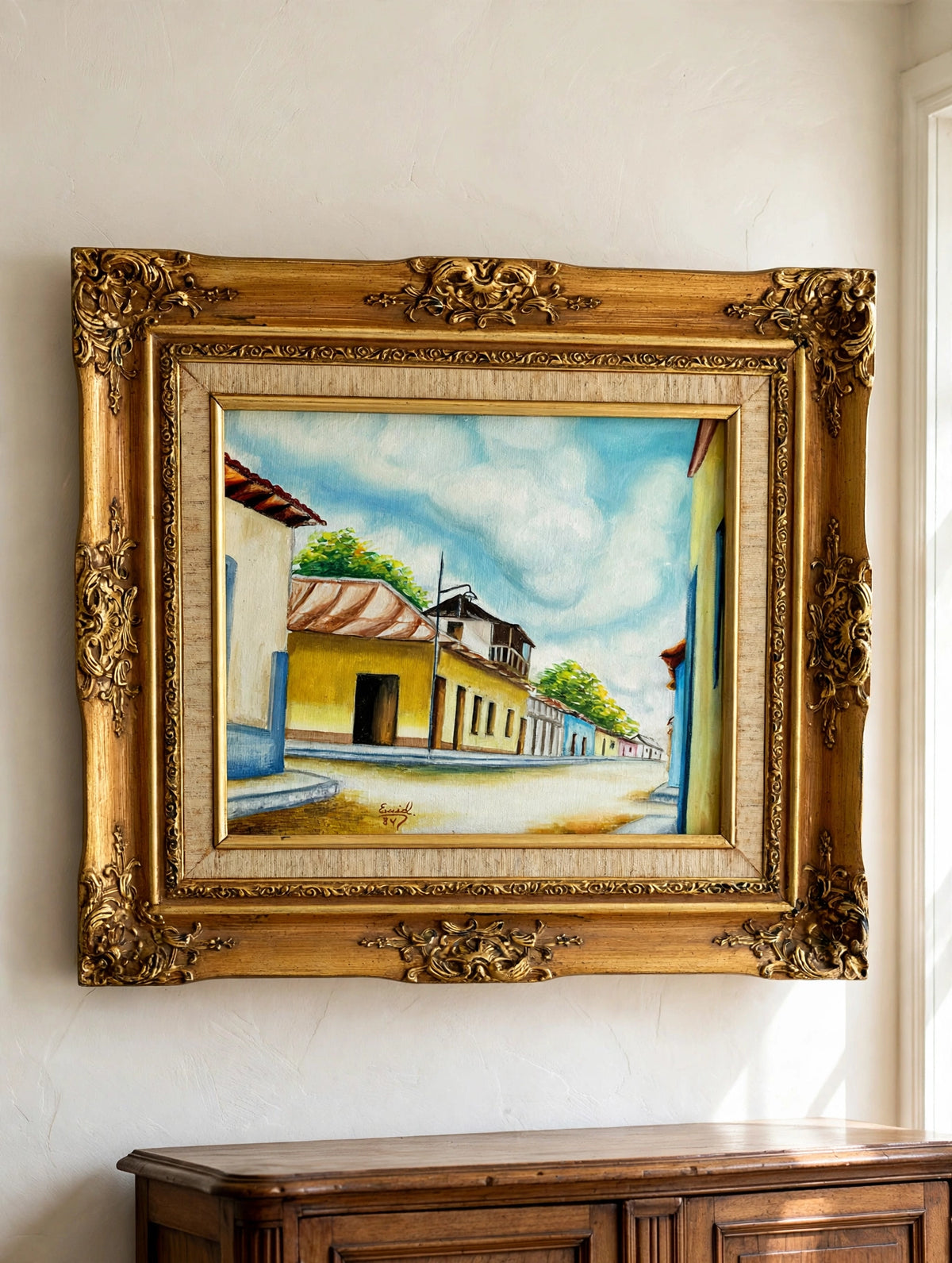Styled lifestyle photo — the 1984 signed Venezuelan street scene oil shown hanging above an antique sideboard in a vintage living room (display inspiration)